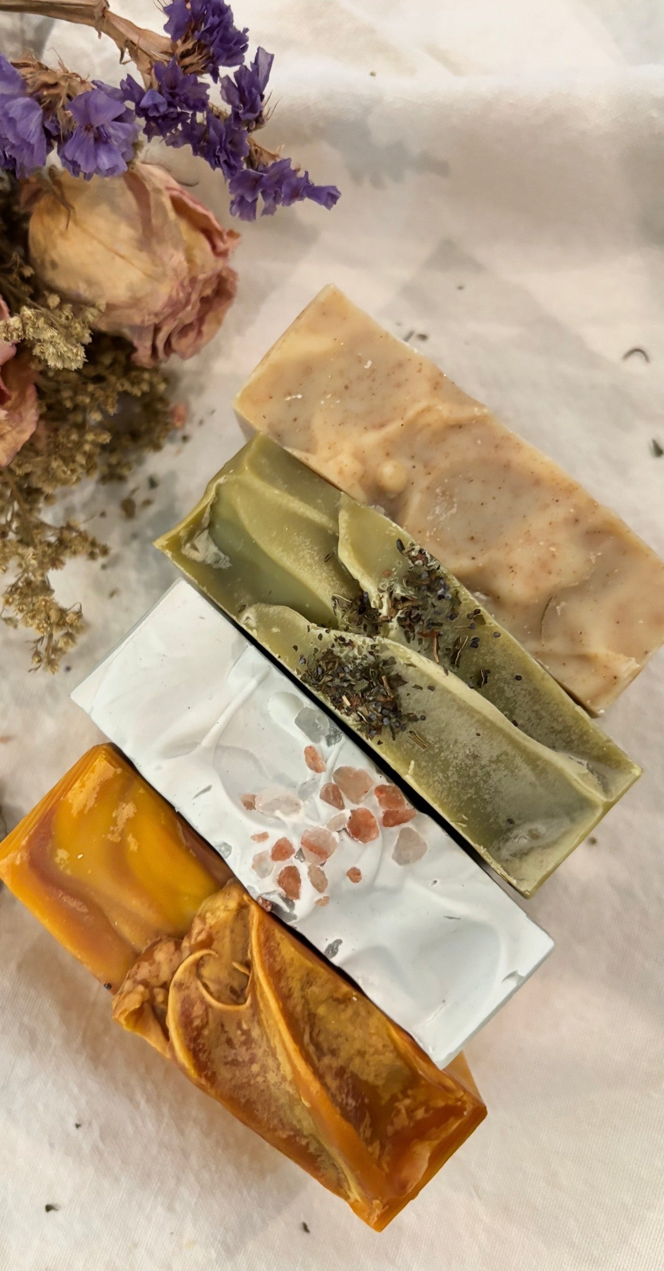 Three handmade soap bars with natural ingredients on a textured surface with dried flowers.